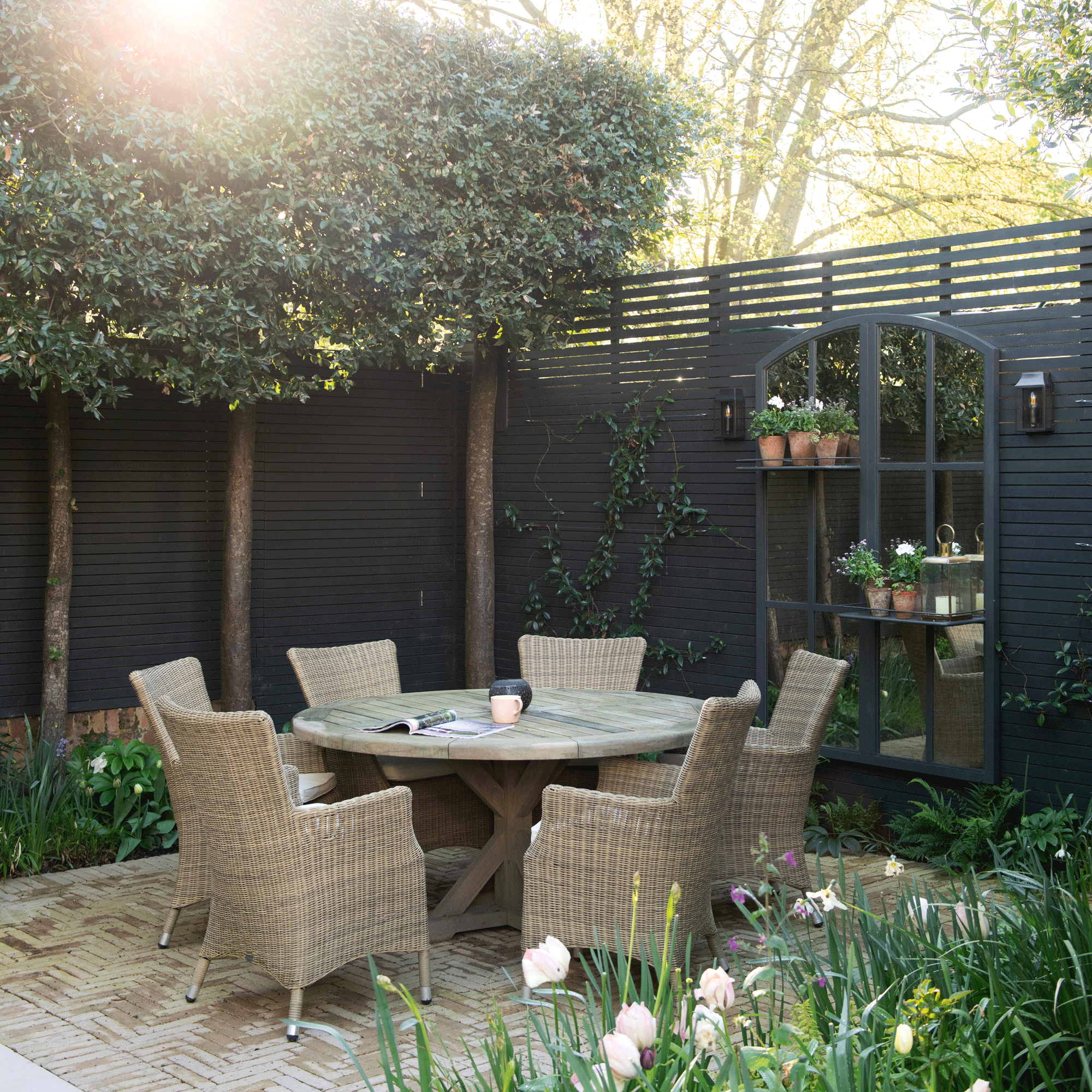 a corner of a garden with black fencing and pleached trees, a dining set and garden mirror
