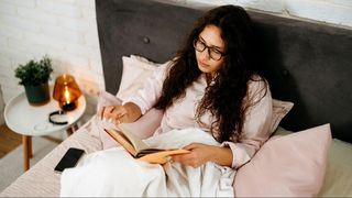 A woman with long dark curly hair and pink pyjamas sitting in bed reading a book