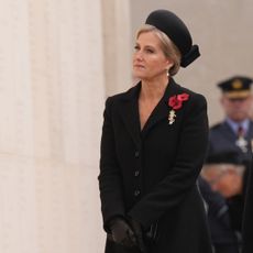 Sophie, the Duchess of Edinburgh wearing a black coat and hat and red poppy brooches standing in front of a war memorial wall