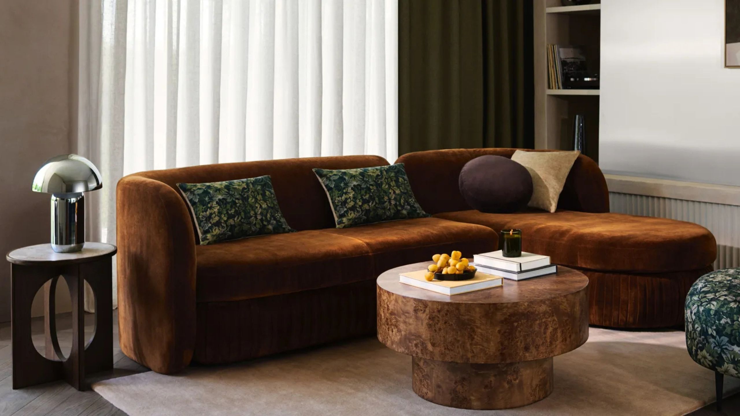 A living room with a modern pleat sofa in a rich brown hue with a mixture of scatter cushions and a round burl coffee table in the center and next to a modern side table and steel mushroom lamp