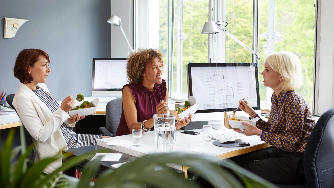 A business owner has lunch at her desk at the office with two of her employees.