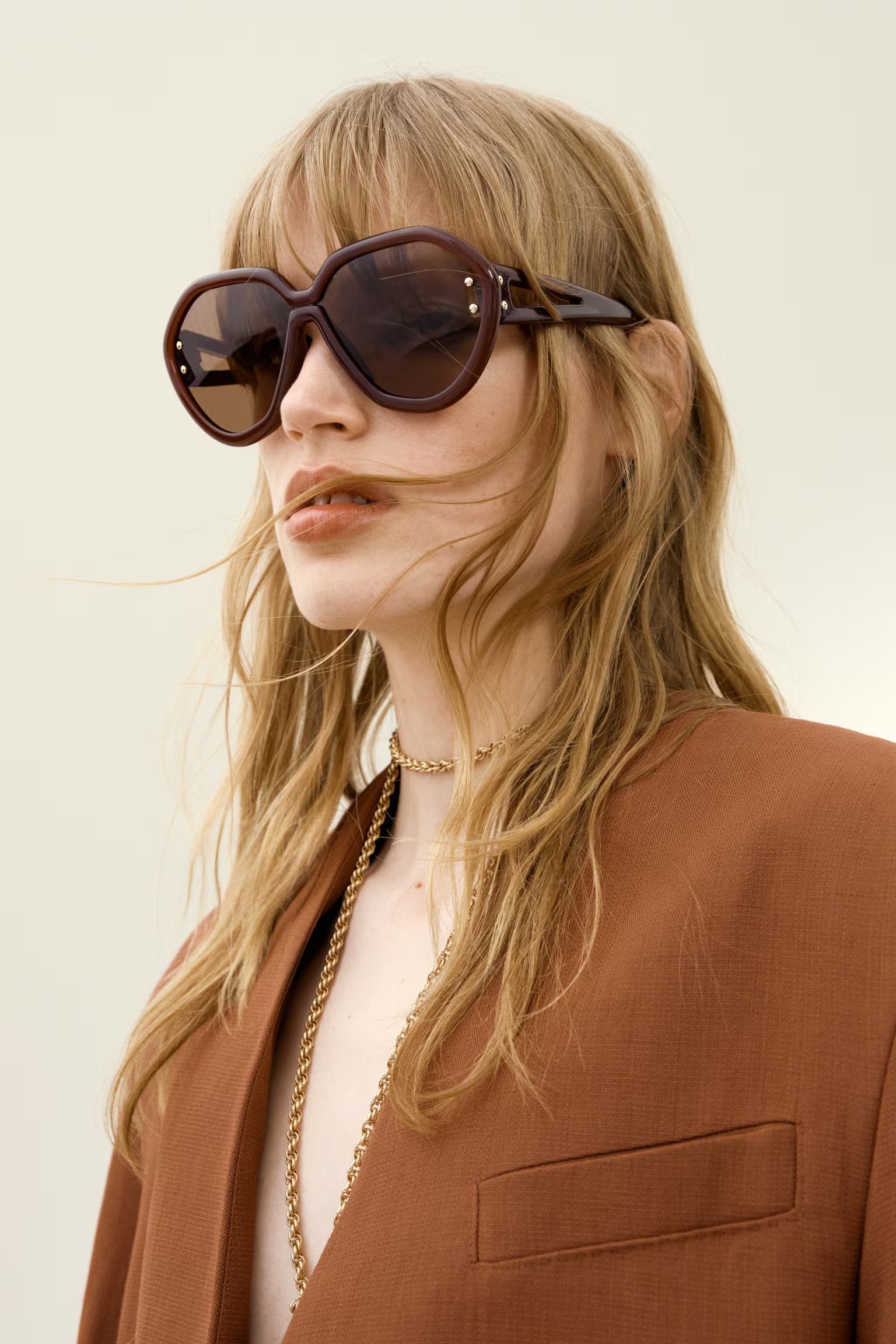 Women wears oversized brown sunglasses, brown blazer and gold necklace