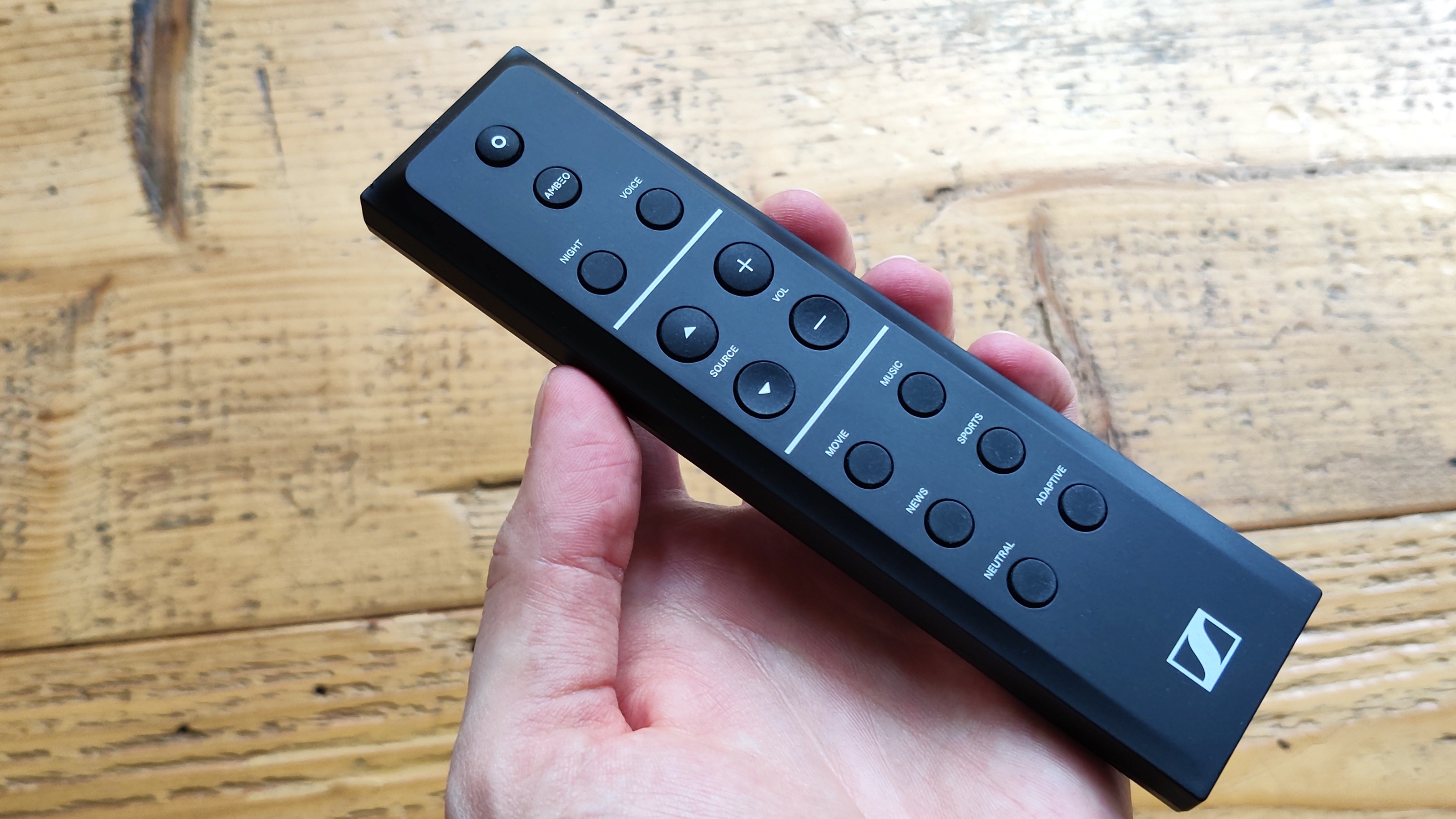 The Sennheiser Ambeo Soundbar Plus remote held in someone's hand above a wooden surface