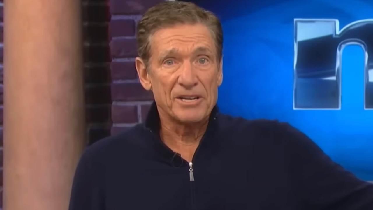Maury sitting on his stage and introducing a guest. He's wearing a black zip up. 