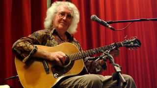 Wizz Jones playing acoustic guitar against a red background
