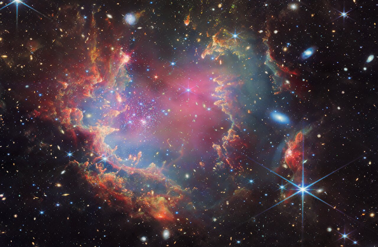 A star cluster is shown inside a large nebula of many-coloured gas and dust. The material forms dark ridges and peaks of gas and dust surrounding the cluster, lit on the inner side, while layers of diffuse, translucent clouds blanket over them. Around and within the gas, a huge number of distant galaxies can be seen, some quite large, as well as a few stars nearer to us which are very large and bright