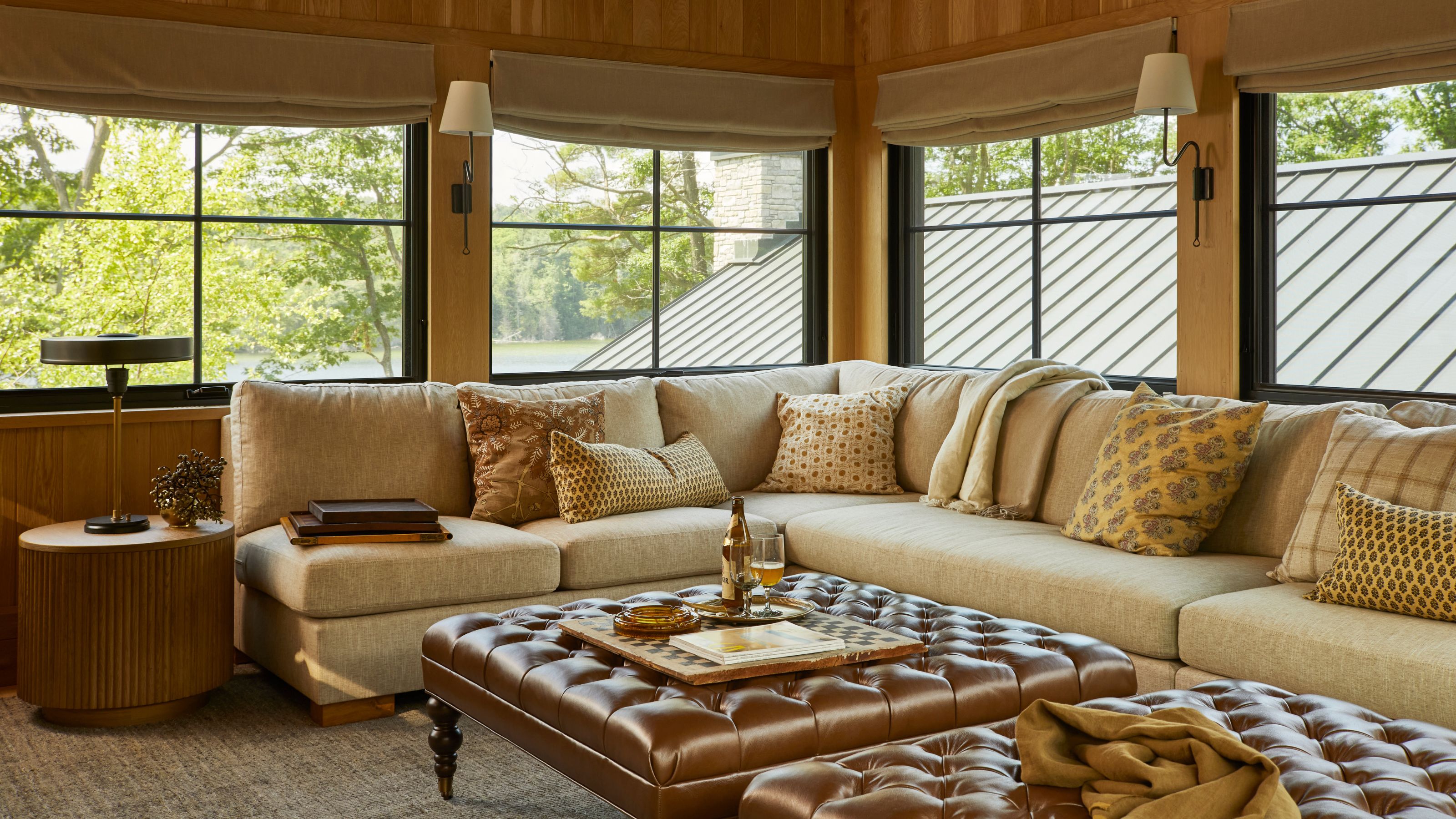 A large, L-shaped sectional sofa in a sunroom wrapped in large windows, featuring a tufted leather ottoman in the center.
