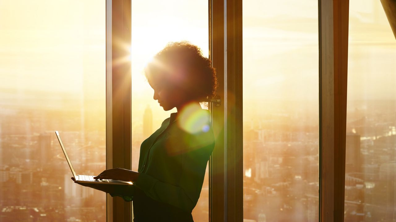 A woman stands at the window of a skyscraper while the sun rises and works on her laptop.