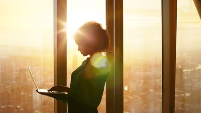 A woman stands at the window of a skyscraper while the sun rises and works on her laptop.