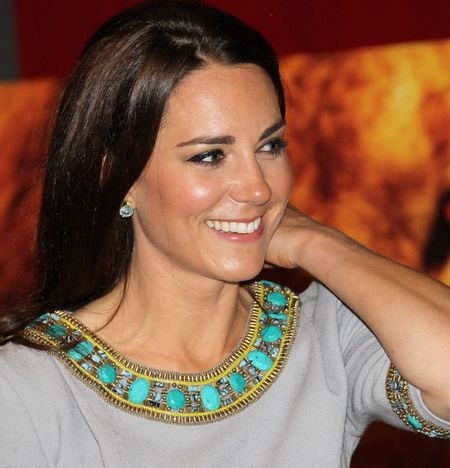 Kate Middleton wearing a gray dress with turquoise details on the neck smiling and resting her arm on her shoulder with a photo of a lion behind her in 2012