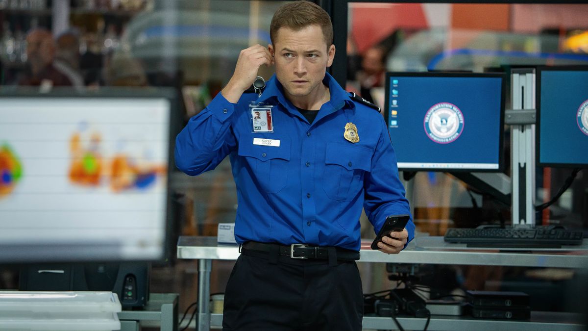 Taron Egerton stands looking confused as he puts an ear piece in Carry-On.