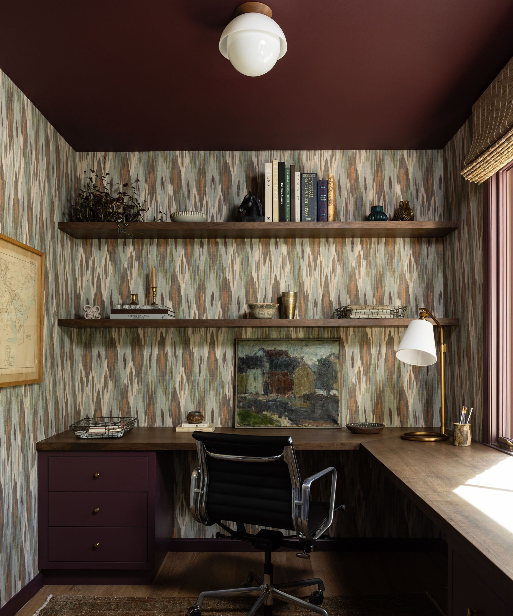 Study with patterned wallpaper, a plum ceiling, an L-shaped wood desk, open shelves and a black leather office chair.