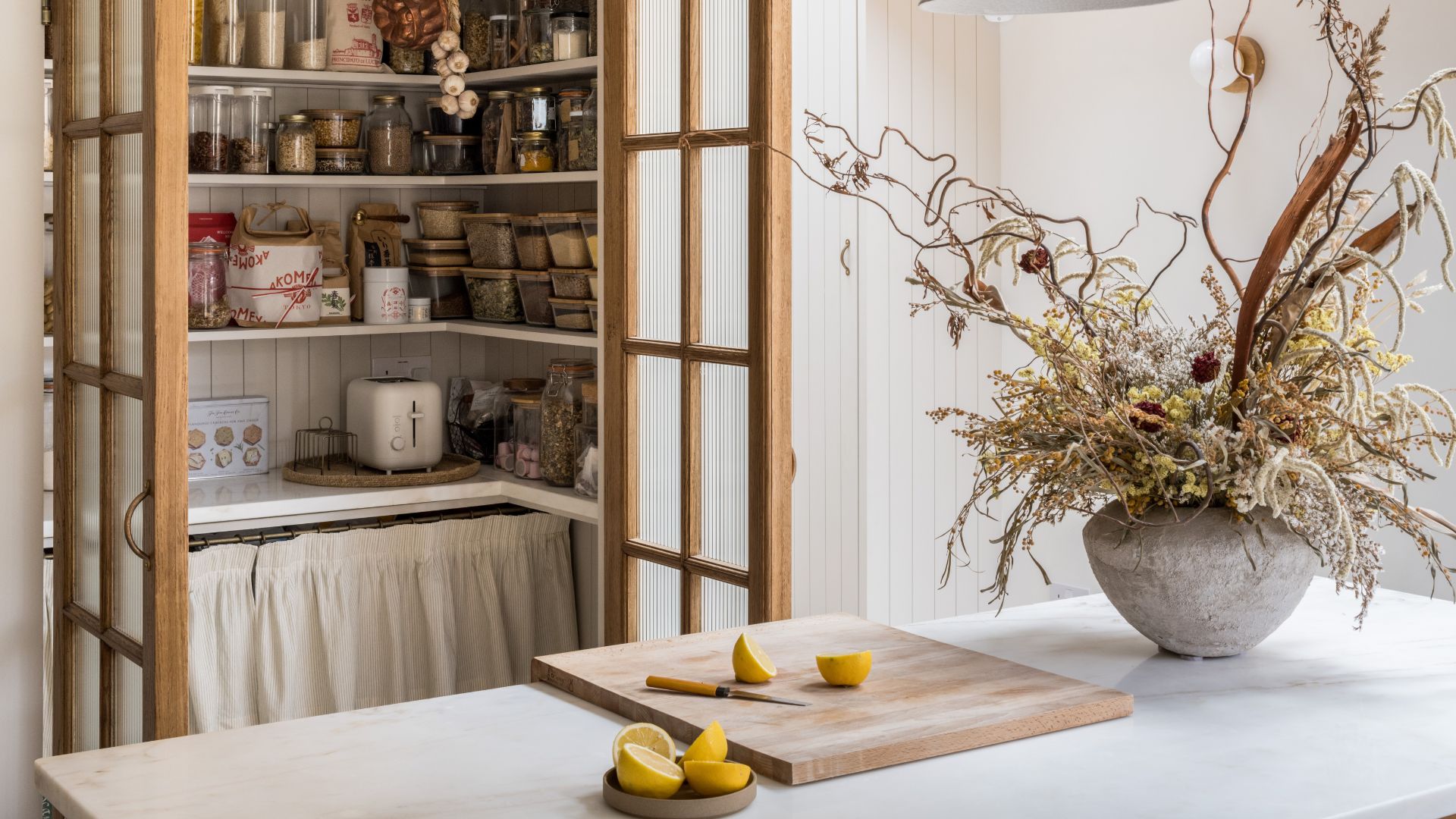 A stone island with a wood chopping board a bowl of limes and a knife near a vase of dried flowers, in front of an open pantry stocked with glass jars