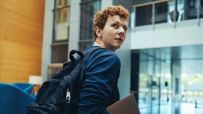 College student carrying his bag and laptop in campus. Young man turning back over his shoulder and walking in college campus.