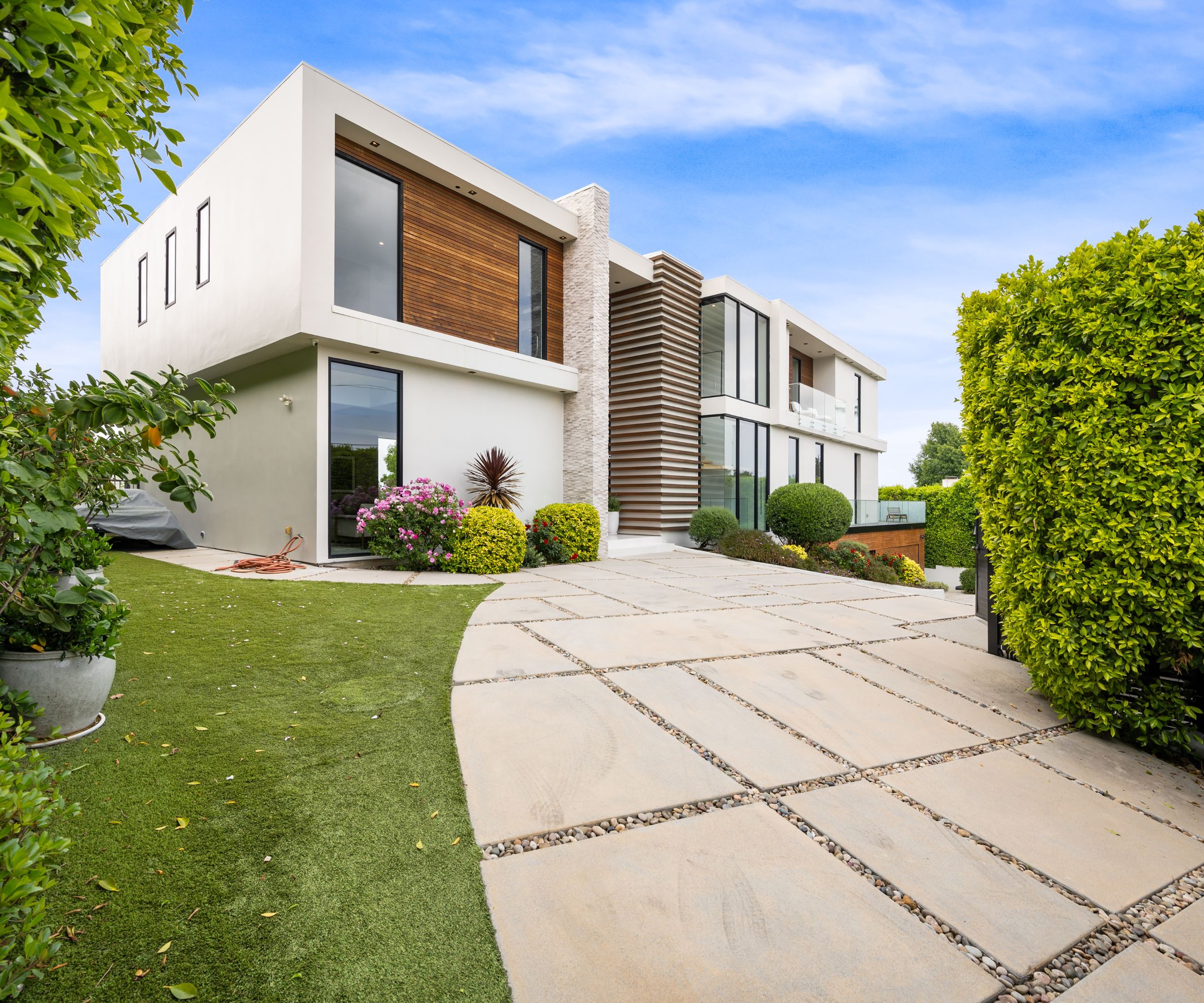 Contemporary two-storey home with white walls, wood and stone accents, and floor-to-ceiling windows. The curved driveway is made of large concrete slabs spaced with gravel joints, bordered by manicured hedges and lush artificial grass.