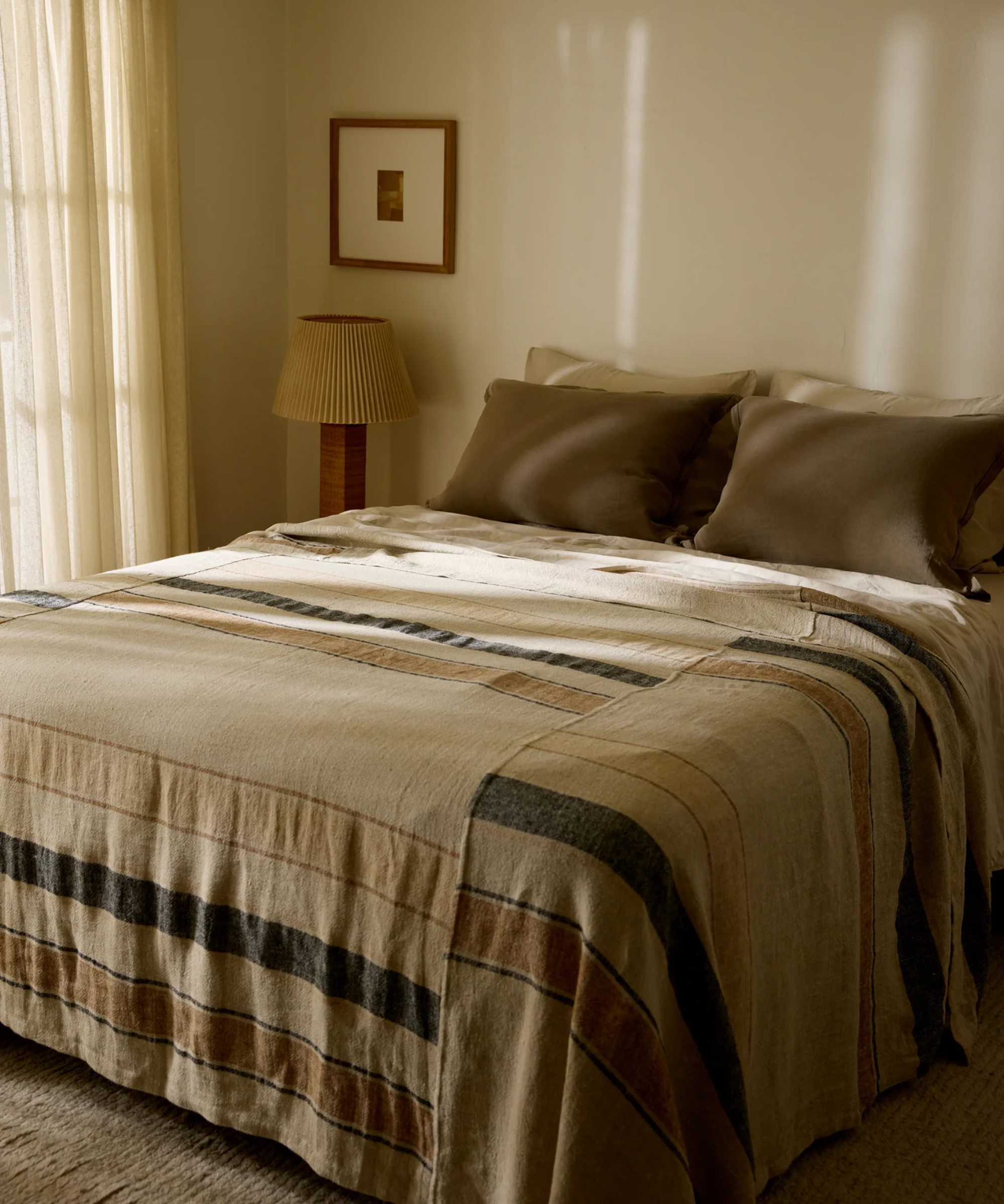 Mocha brown and striped bed sheets pictured in a warm, minimalist bedroom with a pleated table lamp and undersized wall art