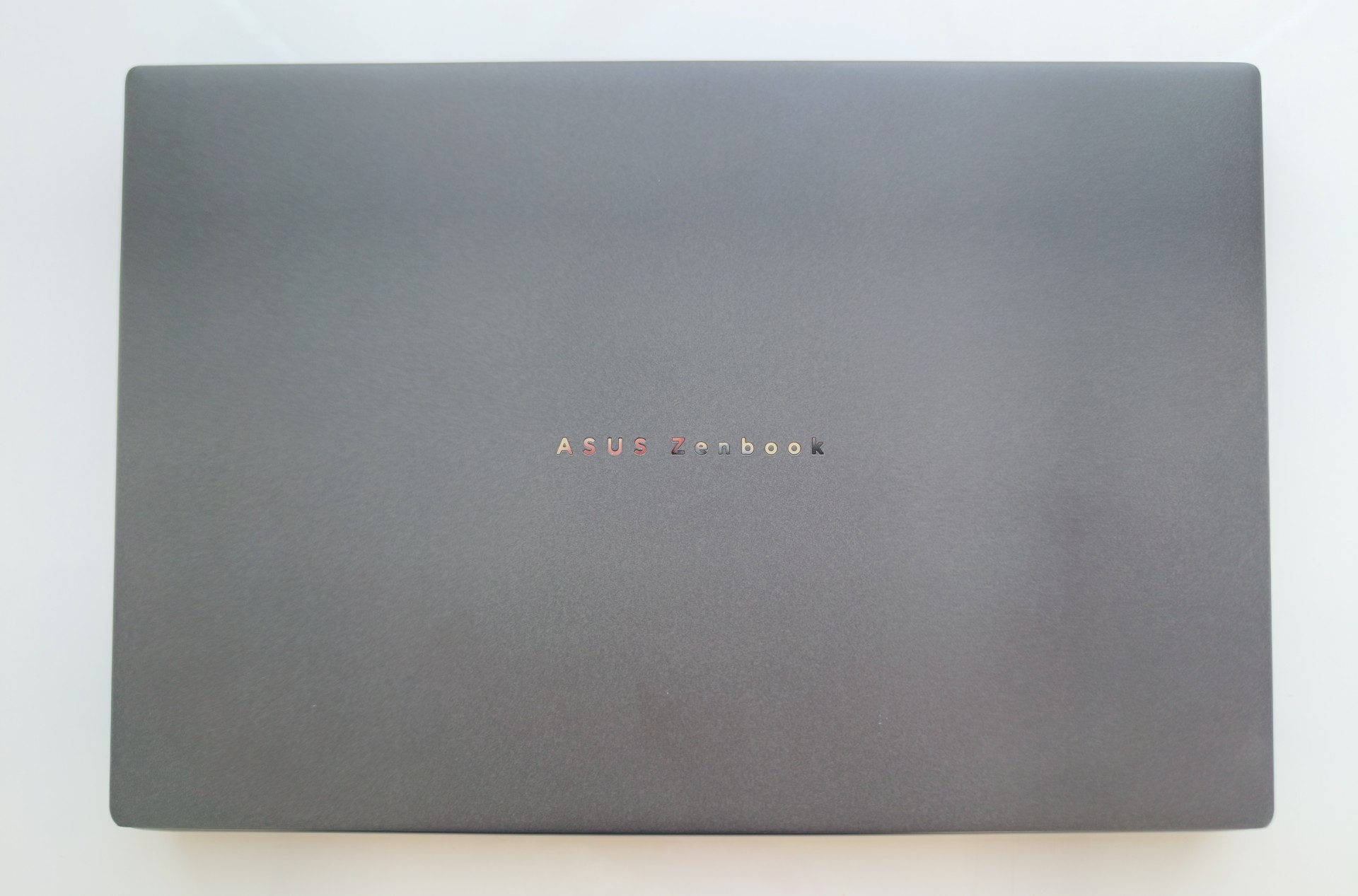 A close-up, top-down view of the dark charcoal grey outer lid of the closed ASUS Zenbook Duo laptop, centered around the sleek "ASUS Zenbook" logo text.