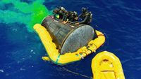 divers in black wetsuits surround a cone-shaped spacecraft in the water being held afloat by yellow floatation devices