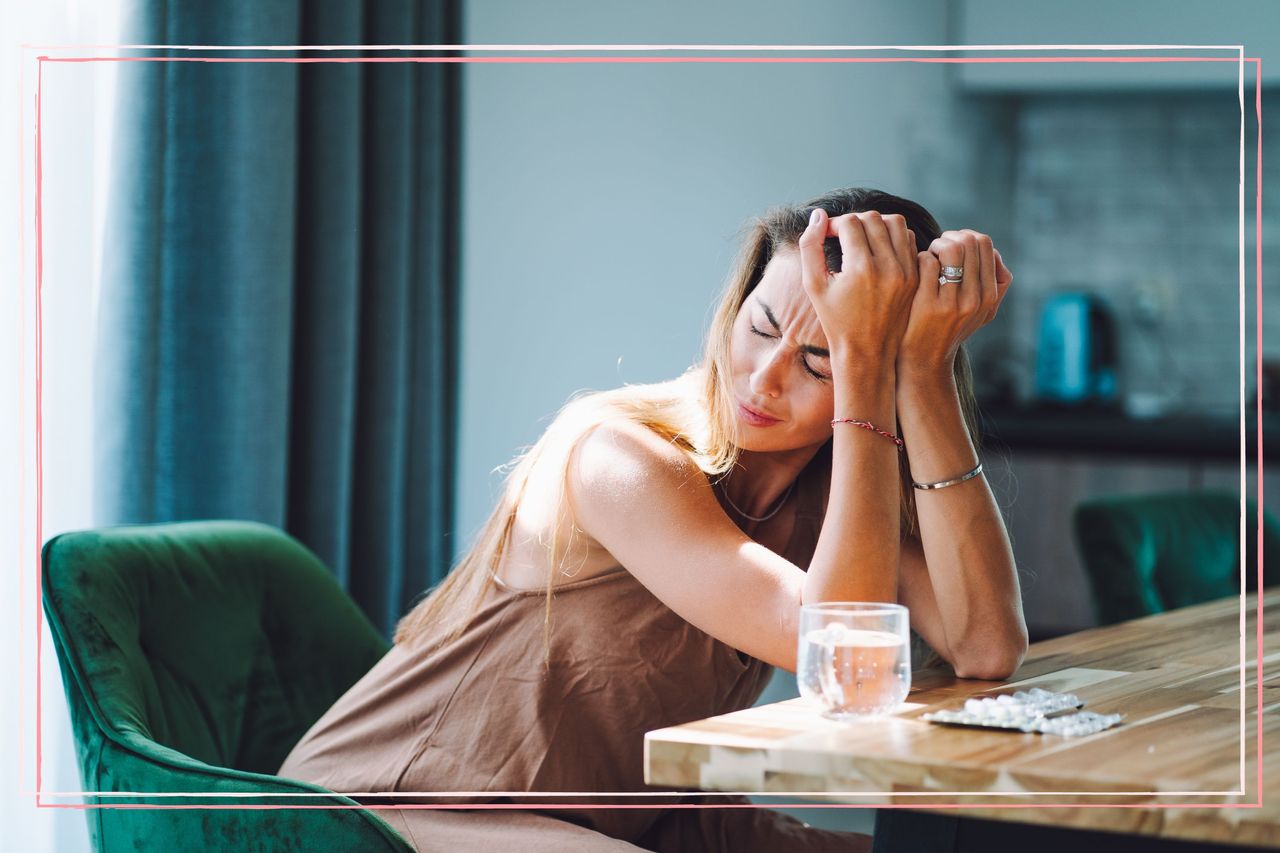 A woman holding her head in her hands next to a glass of water and a packet of painkillers