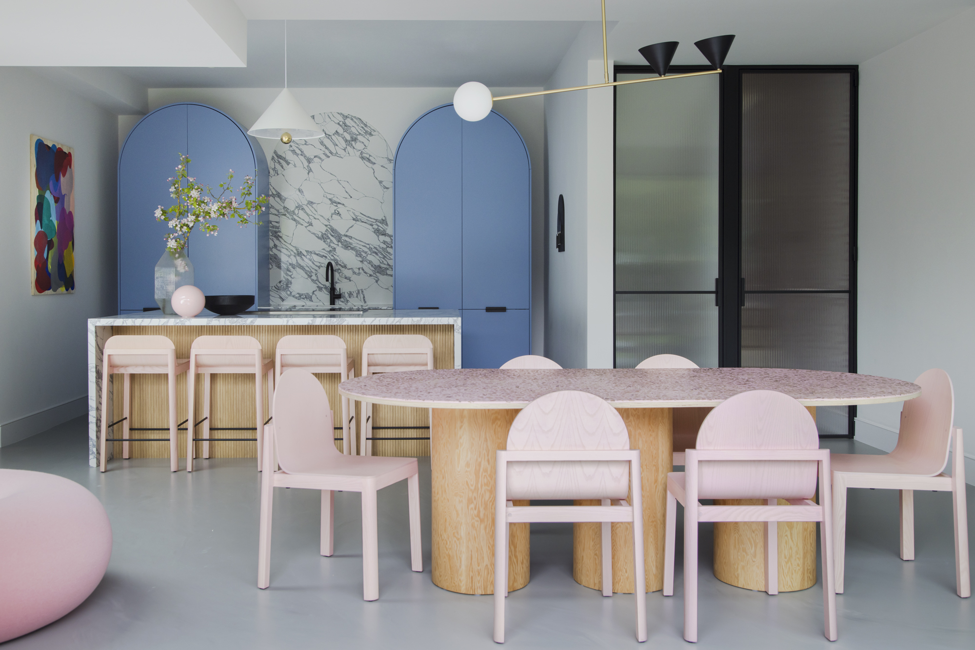 colorful kitchen with curved blue cabinets, marble backsplash and countertop with timber kitchen island, pink bar stool, gray floor, with timber and stone dining table with pink dining chairs and fluted glass doors leading to the hallway