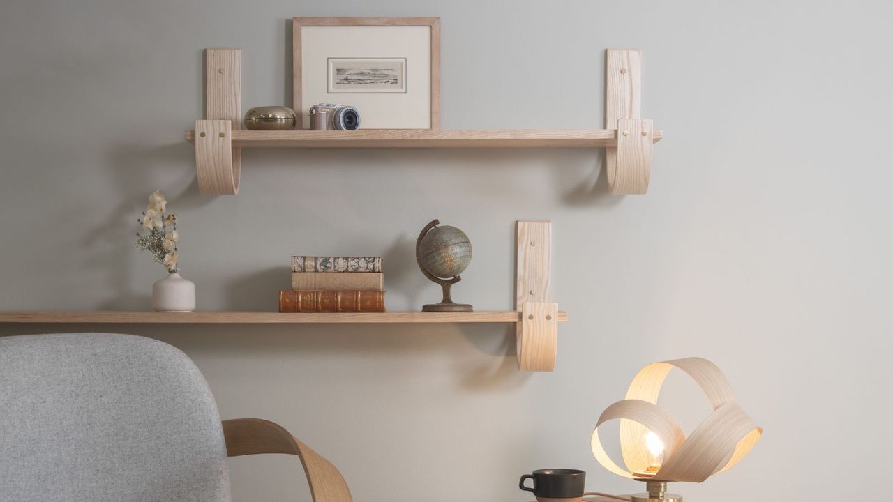 Open wooden wall shelving hanging on a white painted wall. The top of a chair visible in the bottom left corner, and a structural wooden lamp in the right corner. 