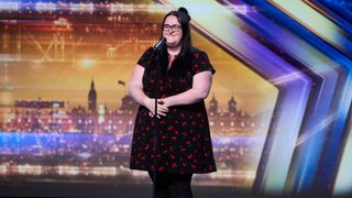 Stacey Leadbeatter in a black dress with red cherries on it, singing into a mic on stage