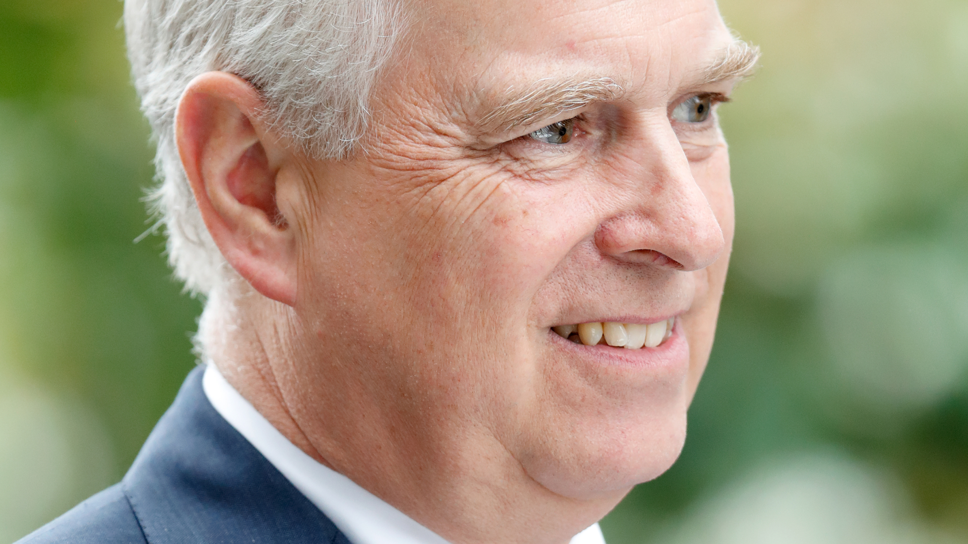 Ex-Prince Andrew wears a navy suit with a white shirt and a red patterned tie and smiles, he has white gray hair