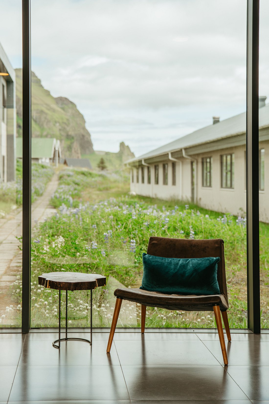 A velvet brown armchair positioned next to a small wood coffee table in front of a panoramic floor-to-ceiling window letting people take in leafy surroundings filled with purple flowers and grass.
