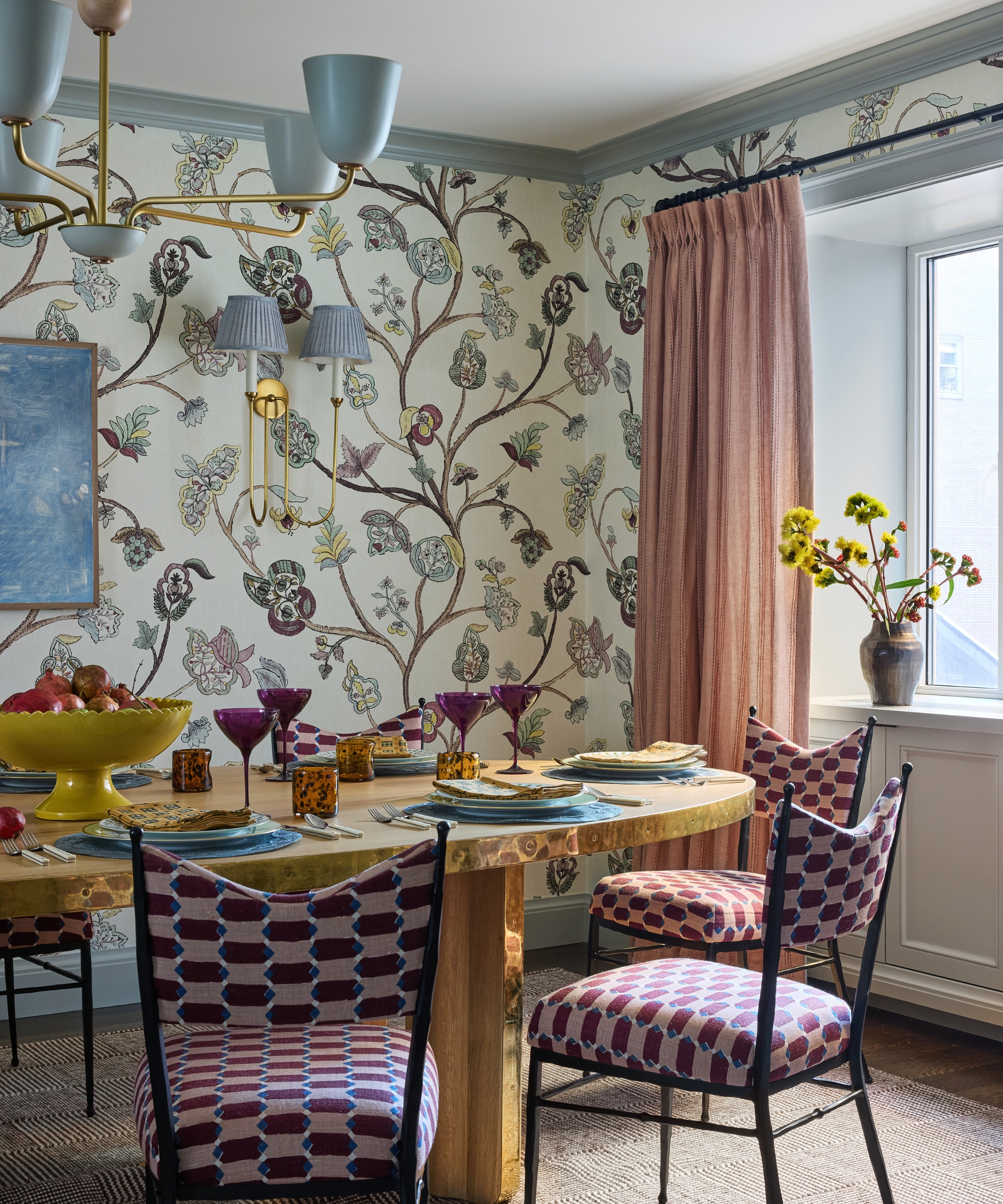 Breakfast room with botanical wallpaper, brass dining table, patterned dining chairs, blue chandelier and blush curtains.