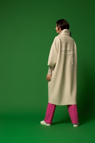 Female model wears long cream coat and pink trousers, against a green background