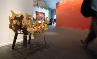 View of the 'Alice' armchair by Philippe Bestenheider - a gold chair featuring multiple 'spinner' type shapes in a space with dark grey flooring and white and red walls