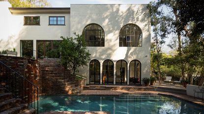 a large LA rendered home with brick patio and stairs with a pool and trees overhead