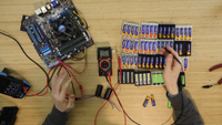 Two hands gesture over 64 AA batteries wired to a volt metre and a stripped down gaming PC.