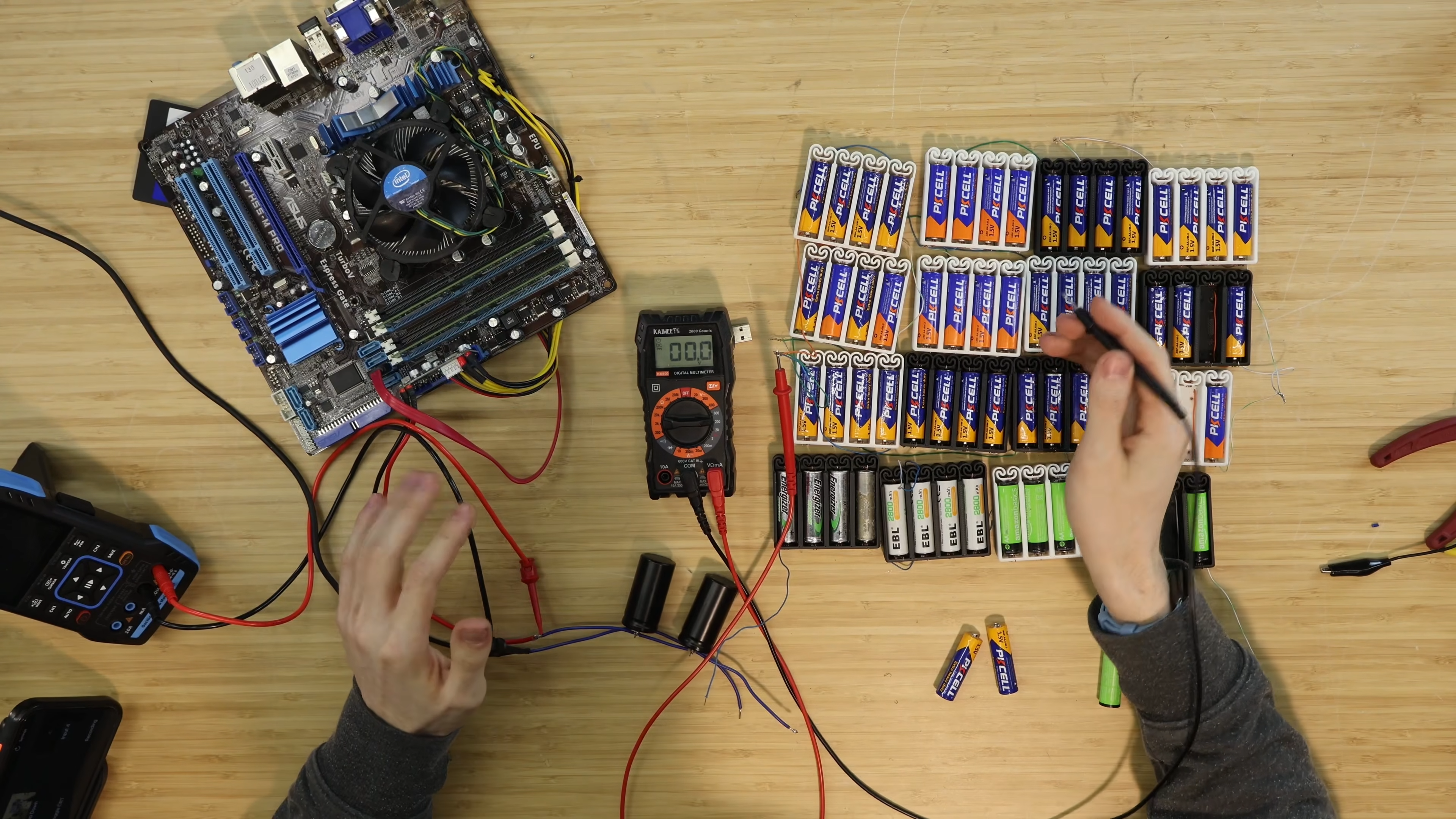 Two hands gesture over 64 AA batteries wired to a volt metre and a stripped down gaming PC.