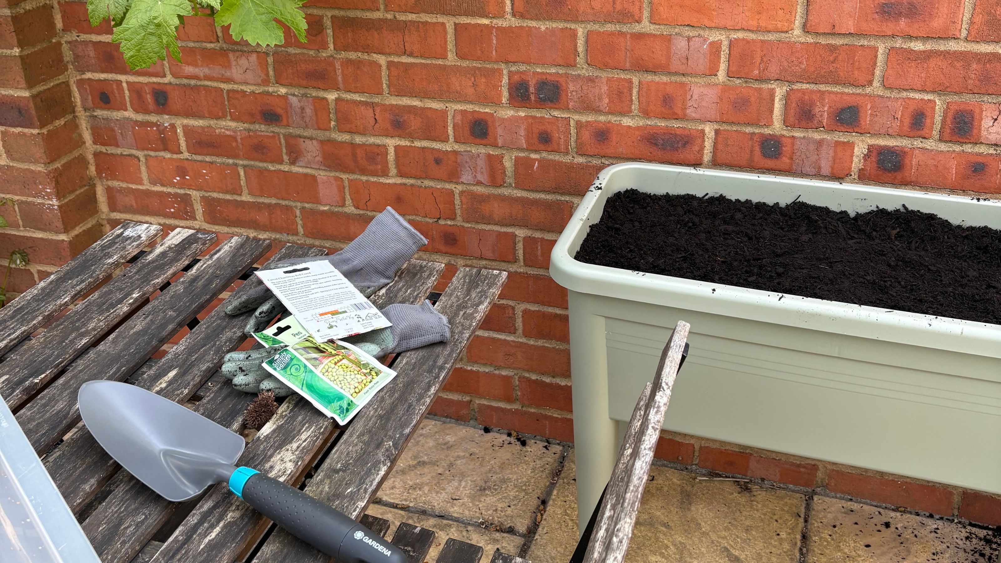 elho Green Basics Grow Table next to wooden garden table and chair, seed packets, garden gloves and GARDENA trowel