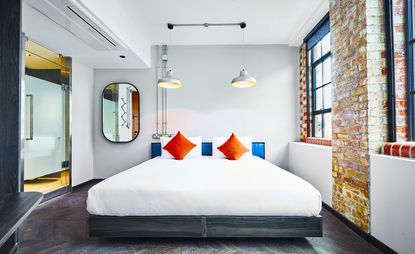 A room in the New Road Hotel. White walls, with an exposed brick section between two tall windows.v Large bed with white linen and bright blue headboard. Mirror on the wall to the left.