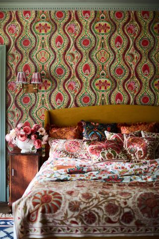 colourful bedroom with paisley wallpaper, mustard upholstered bed, paisley cushions and bedding, wall light, nightstand with vintage vase full of roses, green painted woodwork