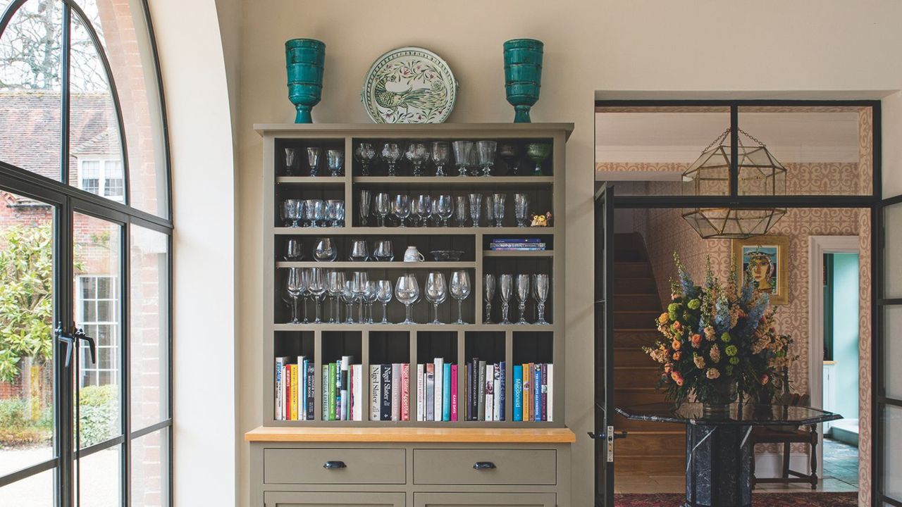 Cabinet with glassware and books, arched window and hallway with large table and flower arrangement