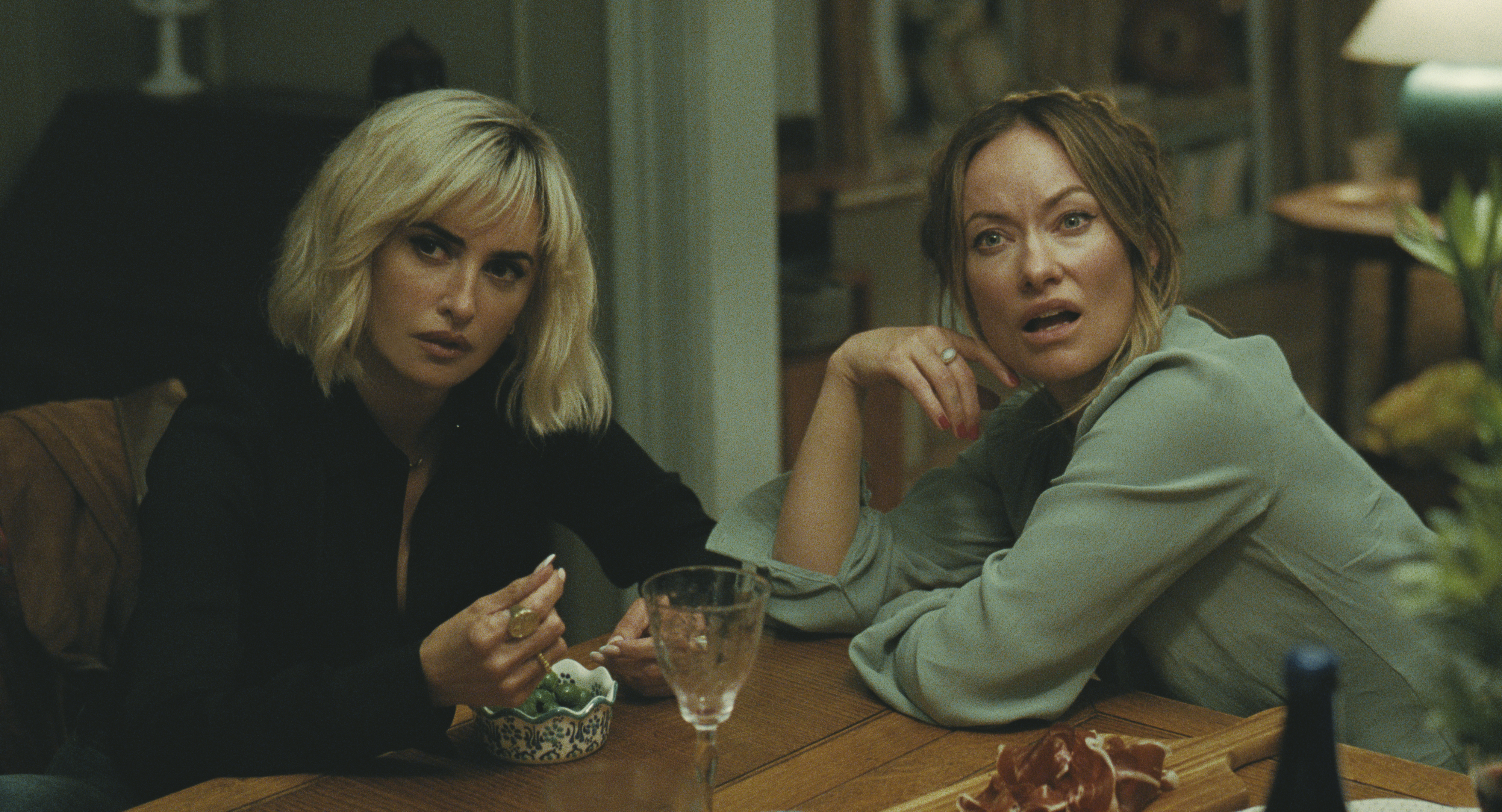 penelope cruz as pina and olivia wilde as angela looking shocked sitting at a dinner table in a still from the invite