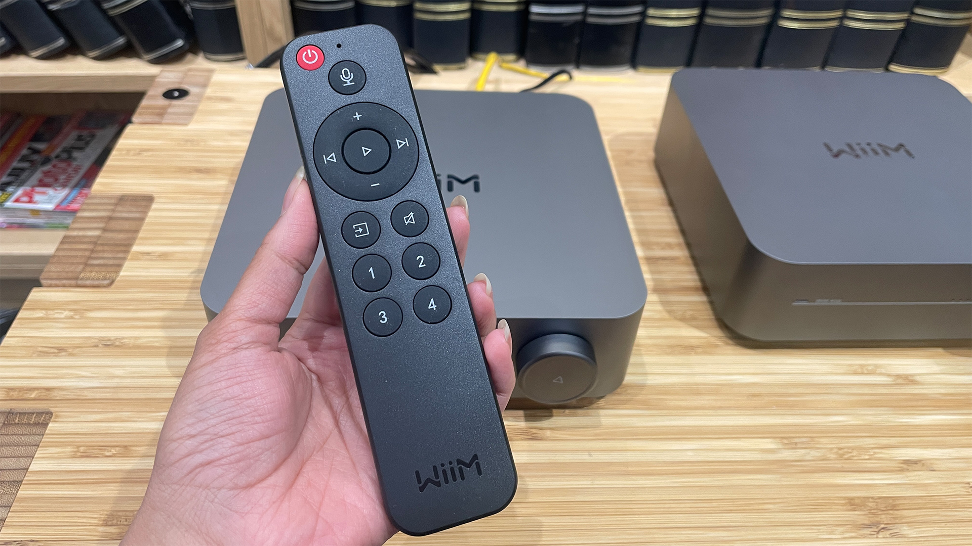 WiiM Amp Pro streaming amplifier remote control held in hand above two WiiM devices