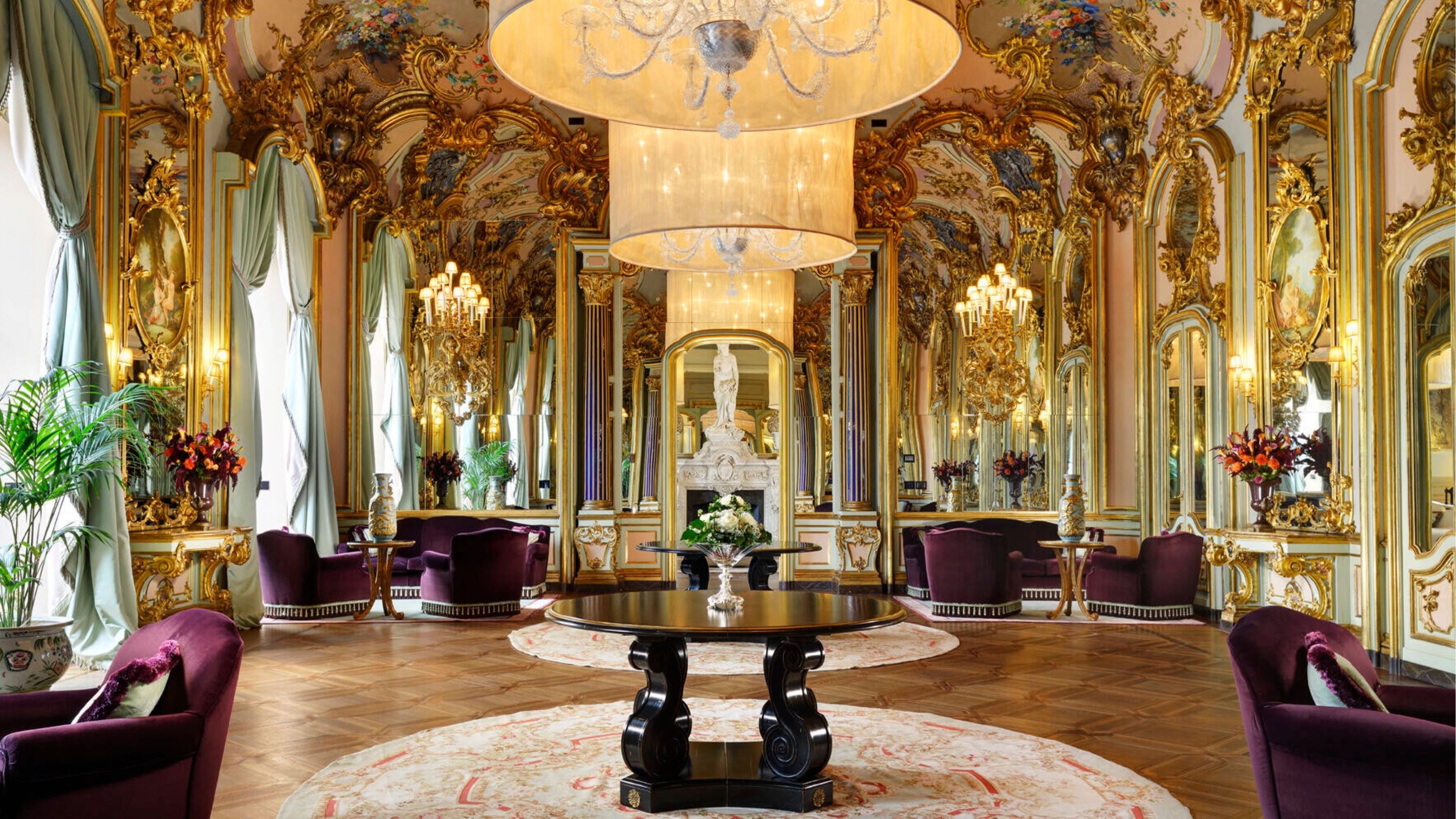 A Baroque hotel lounge characterized by ornate, layered gold leaf details, framed frescoes, mirrors, and large cylindrical pendant lights is furnished with more modern, sculptural armchairs in purple velvet, a central, round wooden table, marble busts, and round orange and cream abstract rugs.