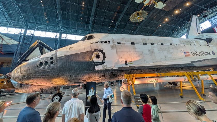 The space shuttle Discovery, a white and black shuttle is on display at the Smithsonian Museum where people walk around it and view it.