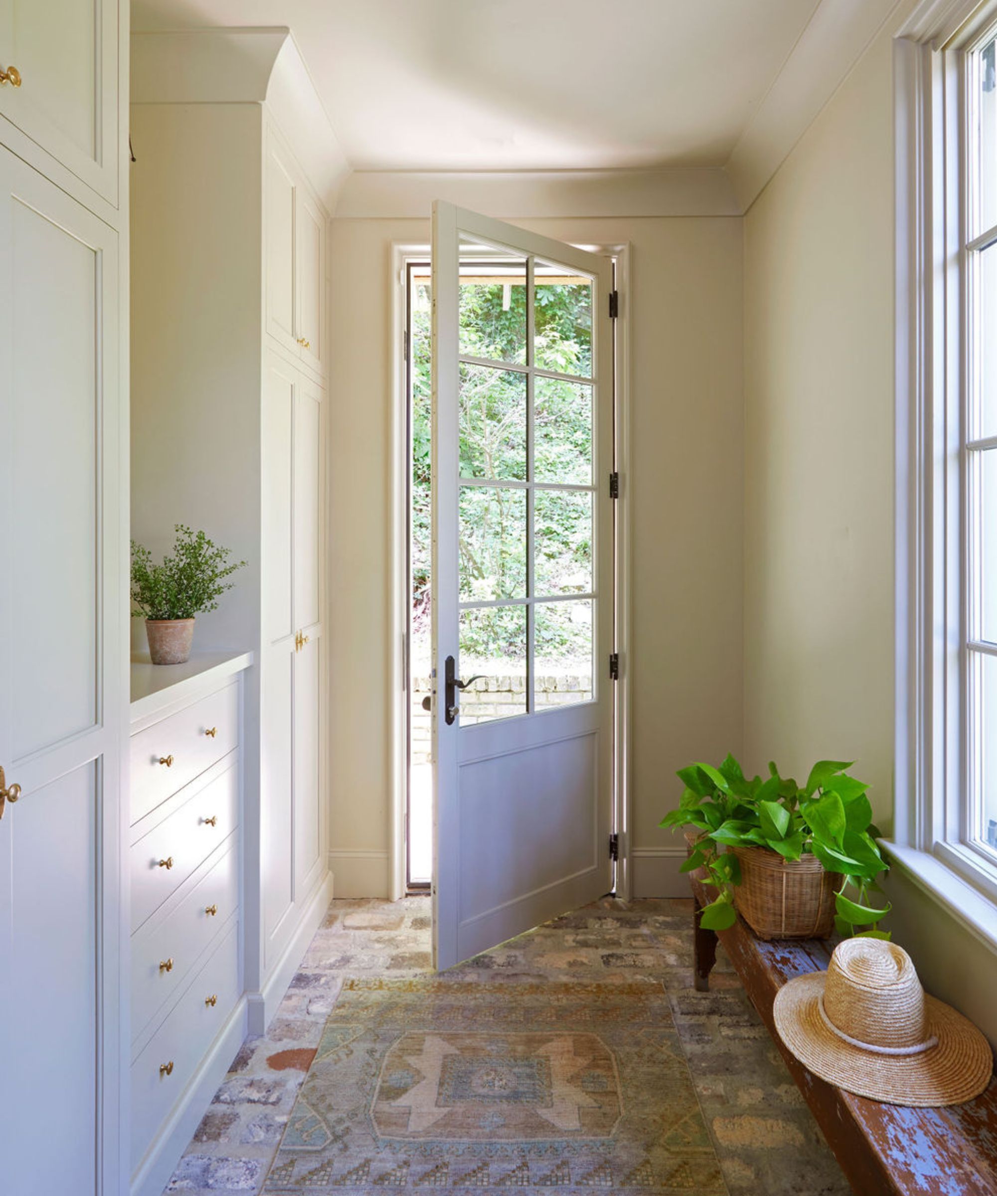 Mudroom with drop cloth by Farrow &amp;amp; Ball painted walls, cream painted cabinetry, stone floor, a wooden rustic bench with a potted plant and a hat on top, and a back door with glass panelling