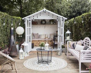 Wildflower Garden Summerhouse, a white summerhouse at the end of decking, with a wooden bench with cushions inside, and outdoor seating, a rug and a black coffee table outside on the decking