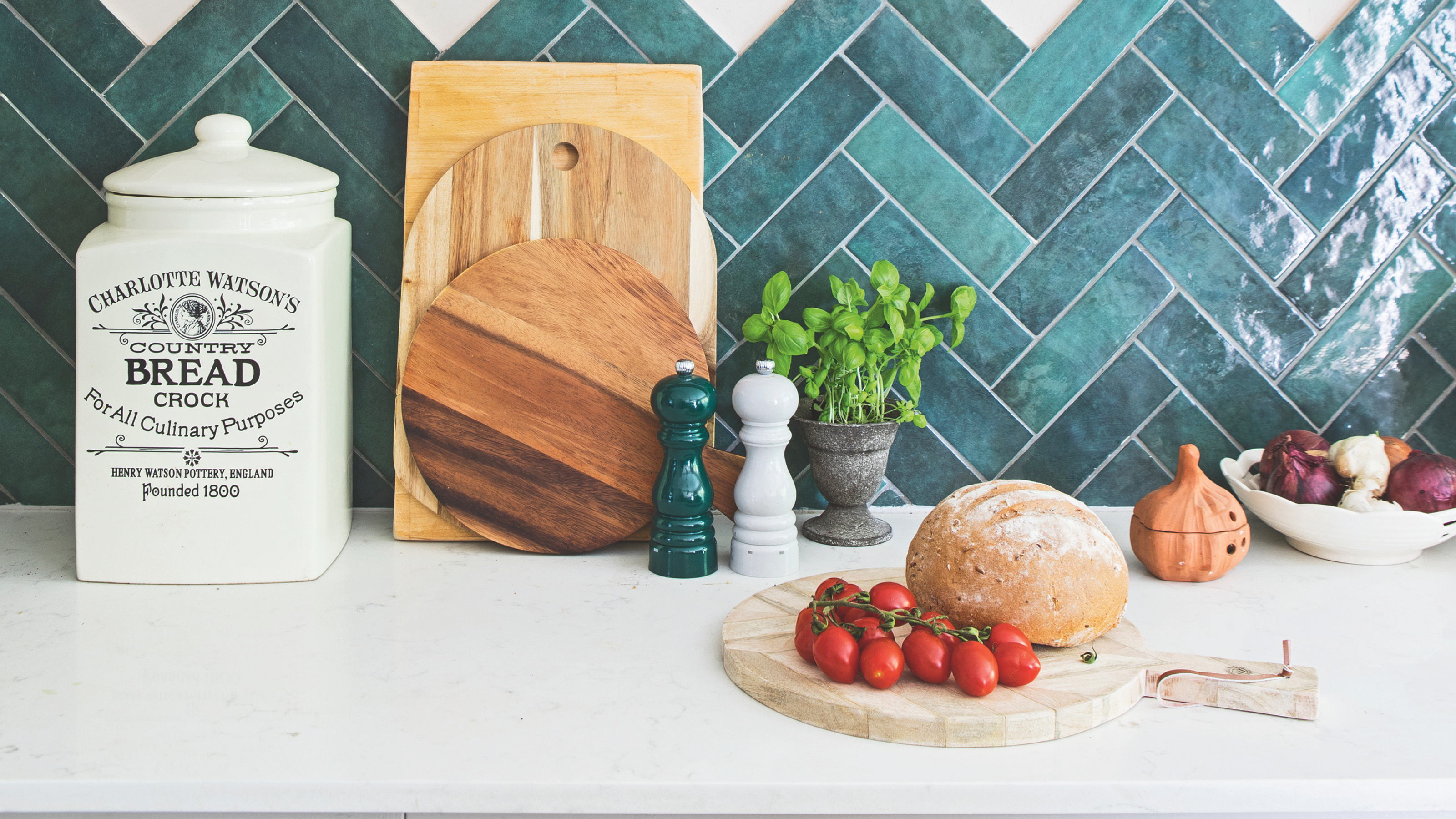 A kitchen work top area with green tiles and white surface. On top are three chopping board stacked against the wall, a vintage bread flour jar, salt and pepper mills, basil plant, salt pot, bowl of onions and board with fresh bread and tomatoes on top.