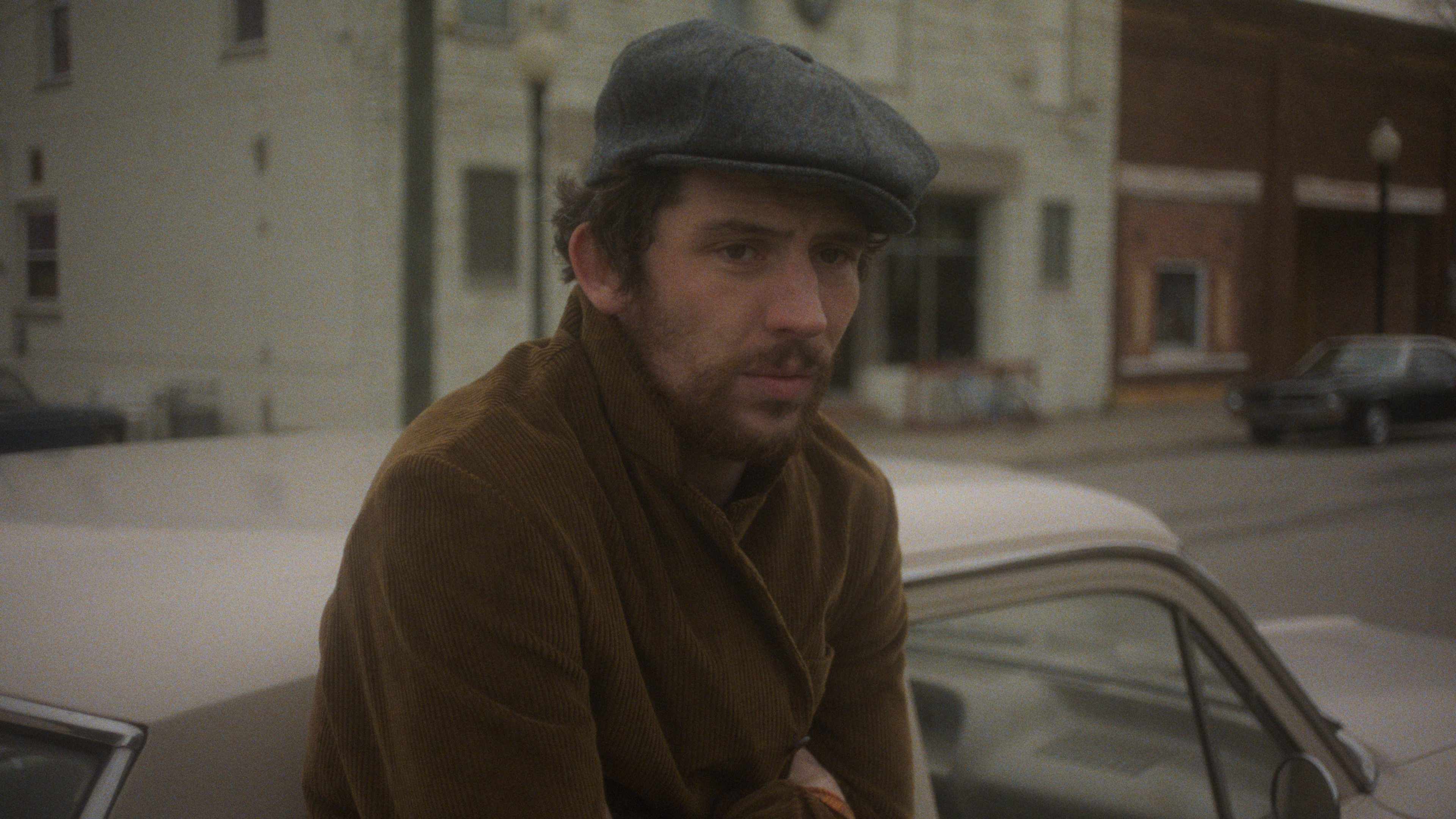 josh oconoor wears a cap and suede jacket while standing next to a car in the movie the mastermind