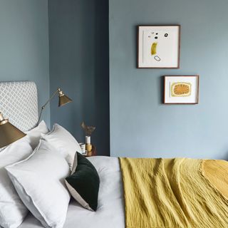Blue bedroom with a mustard throw, white bedding, upholstered headboard, wall art