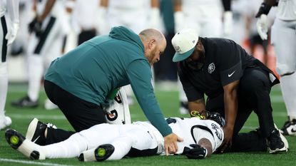 A football player in a white uniform lays on a green field tended two by two men wearing teal and black clothes.
