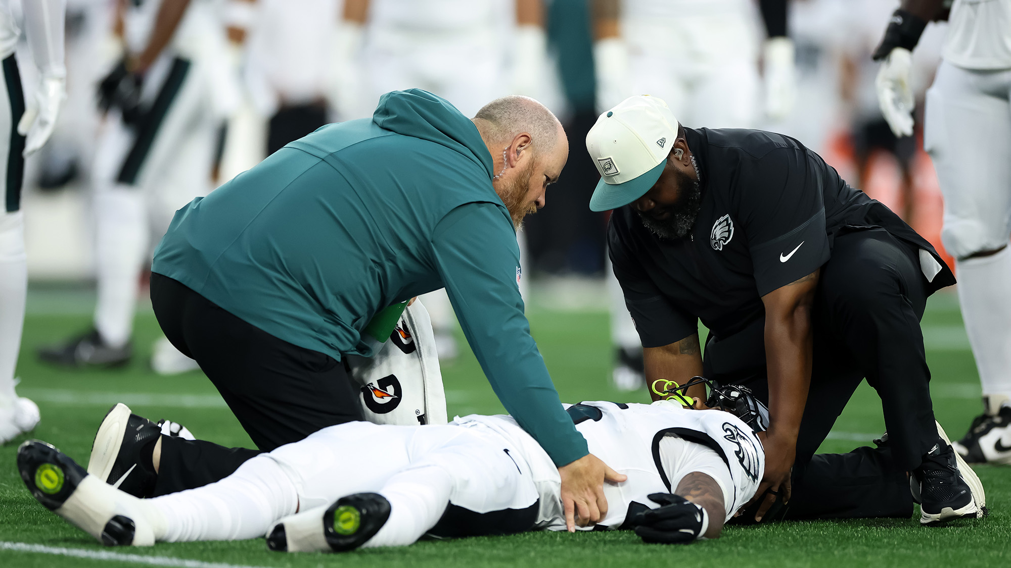 A football player in a white uniform lays on a green field tended two by two men wearing teal and black clothes.