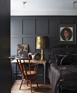 Bedroom with black painted paneling, black metal bedframe with black duvet, wood and metal desk with dark wood traditional armchair, paintings on desk and on walls, metallic lamp shade